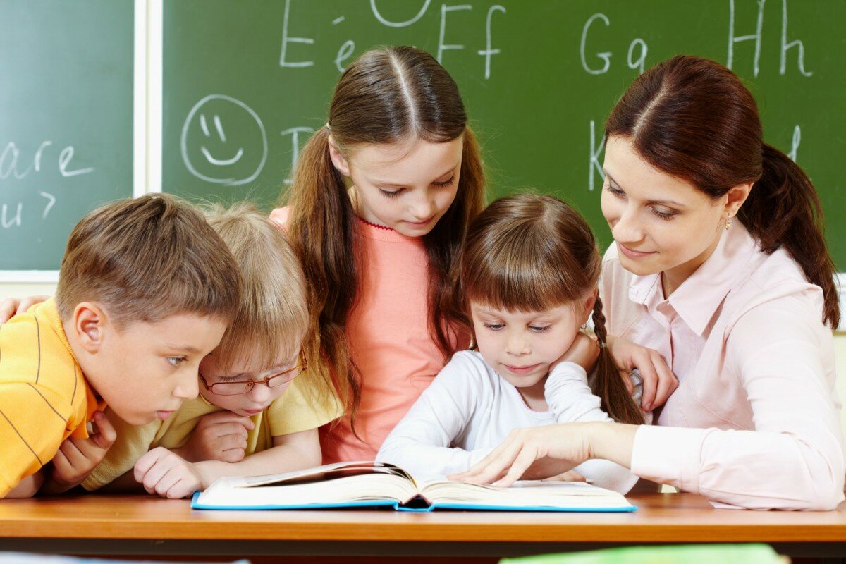 Мальчики и девочки на столе у школьной доски вместе са учителем читают книгу